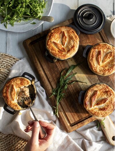 Individual pies with leaf-decorated pastry lids in small black casserole dishes, one opened with a fork