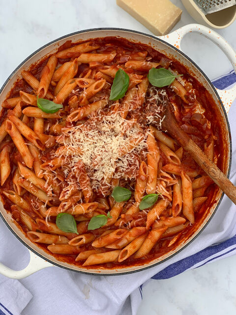 A pan of penne in tomato sauce topped with grated parmesan and basil leaves, with a wooden spoon