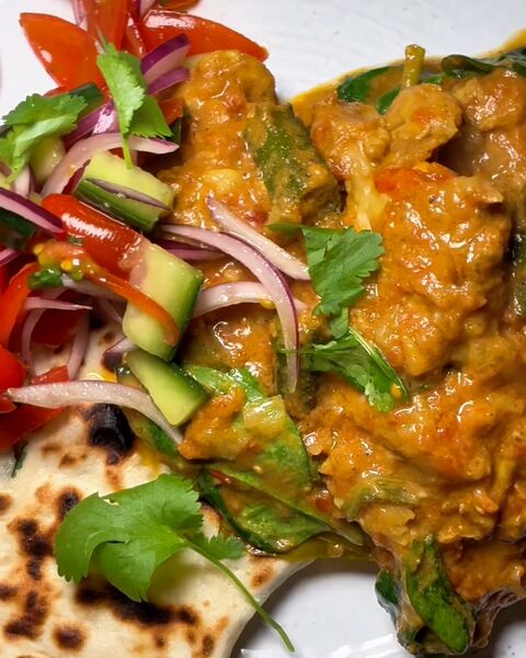 Spiced chicken curry on a white plate with a charred flatbread, topped with red onion, tomato, cucumber and coriander
